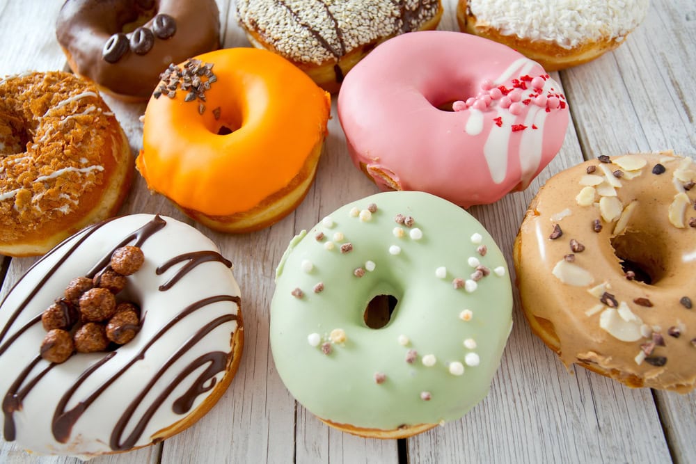 Assorted donuts with colorful icing and various toppings arranged on a white wooden surface.