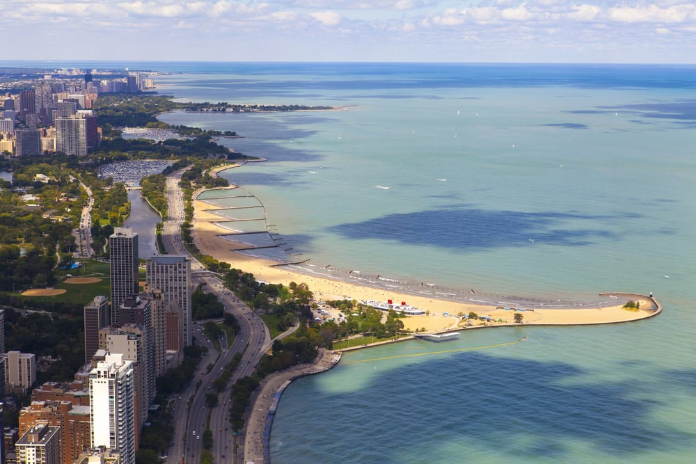 Aerial view of a sandy beach and lakeshore with city buildings, a road, green parks, and calm blue water under a partly cloudy sky.