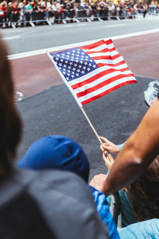 A person in a crowd holds a small American flag on a stick, with a street and more people in the background.