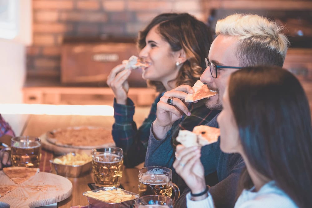 Three people sit at a table, eating slices of pizza and drinking beer or cider, with additional pizza and snacks on the table in front of them.