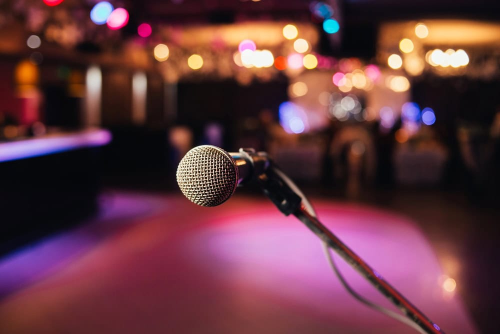 A close-up of a microphone on a stand with a blurred, colorful background showing lights and an indoor venue.