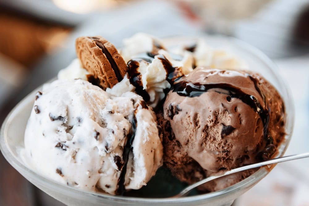A glass bowl with scoops of chocolate and cookies and cream ice cream, topped with whipped cream, chocolate syrup, and a chocolate cookie; a spoon is on the side.