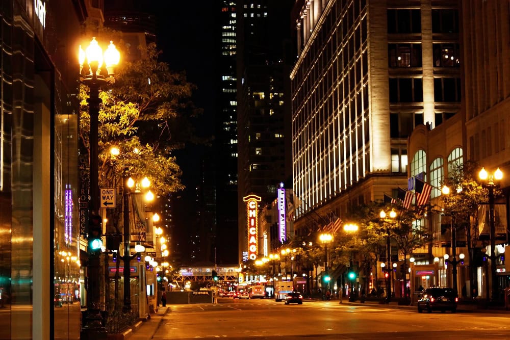 Night view of a well-lit city street with the Chicago Theatre sign visible, lined with buildings, street lamps, and flags.