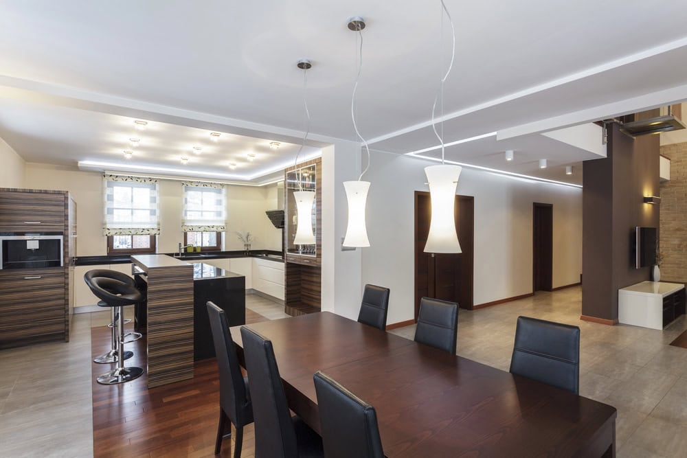 Modern open-plan kitchen and dining area with wooden cabinetry, a dark dining table with black chairs, pendant lights, and neutral colored floors and walls.