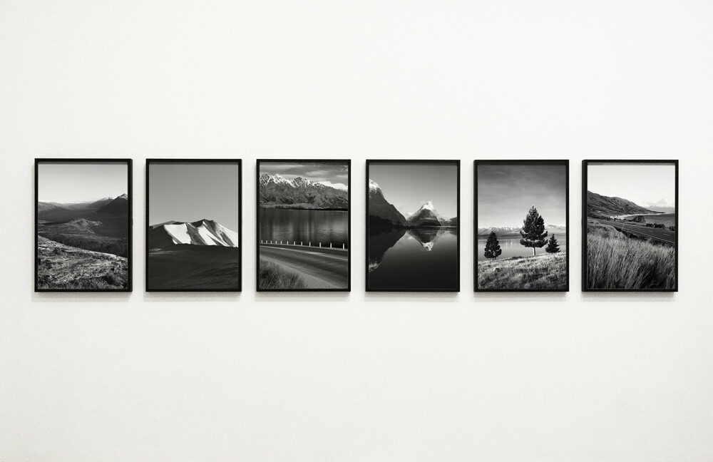 Six black-and-white landscape photographs in black frames are evenly spaced and displayed in a row on a plain white wall.