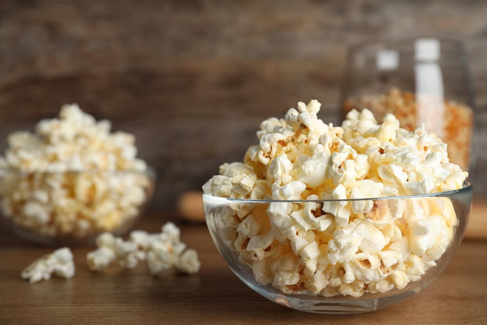 A clear glass bowl filled with popped popcorn sits on a wooden surface, with another bowl of popcorn and a jar of unpopped kernels in the background.