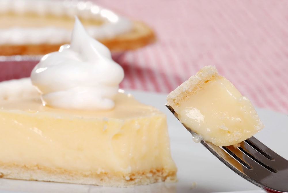 A close-up of a fork holding a bite of creamy pie, with a slice of pie topped with whipped cream on a plate in the background.