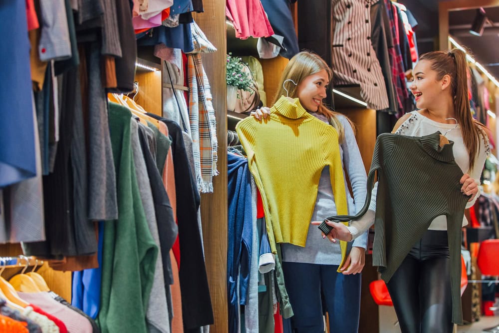 Two women stand in a clothing store, each holding up a sweater and smiling at each other while surrounded by racks of clothes.