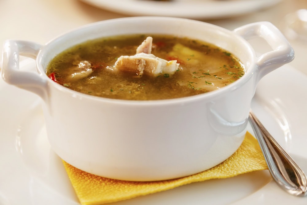 A white ceramic bowl filled with clear chicken soup containing pieces of chicken and vegetables, placed on a yellow napkin with a spoon beside it.