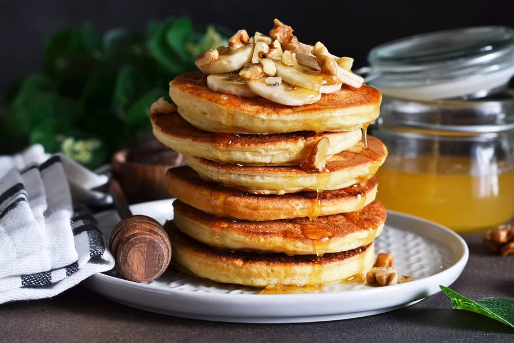 A stack of pancakes topped with banana slices and chopped nuts, drizzled with honey, on a white plate with a honey dipper and jar in the background.