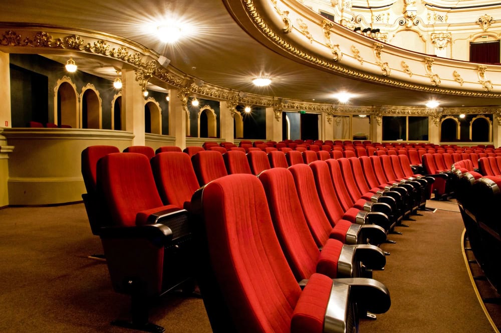 Rows of empty red velvet theater seats face a stage, surrounded by ornate gold architectural details and balcony sections in a classical theater interior.