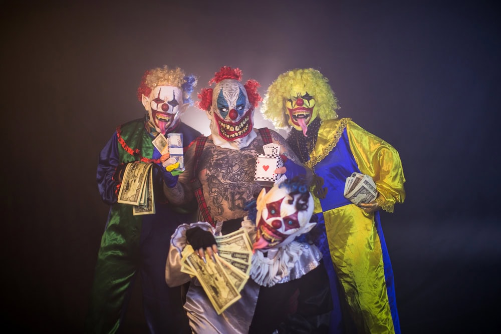 Four people in scary clown costumes and masks pose with playing cards and fanned stacks of cash against a dark, smoky background.
