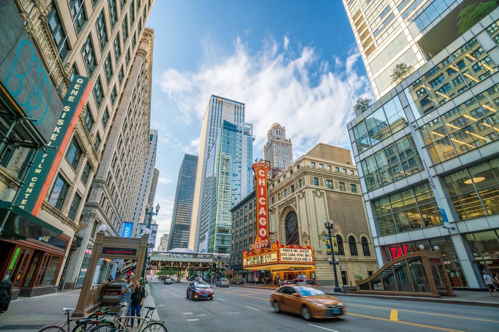 A busy downtown Chicago street with cars, cyclists, pedestrians, tall buildings, and the iconic Chicago Theatre marquee under a partly cloudy sky.