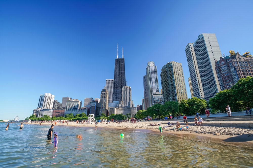 People enjoy a sunny day at a sandy beach with calm water, with a city skyline featuring tall buildings and greenery in the background.