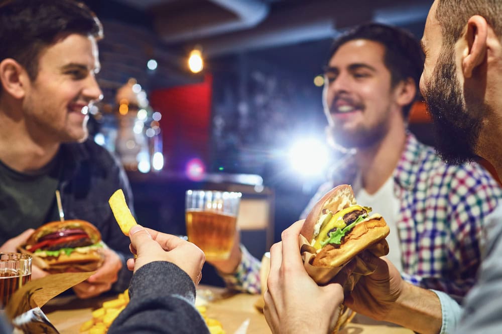 Three men sitting at a table in a restaurant, eating burgers and fries, with drinks and talking together.