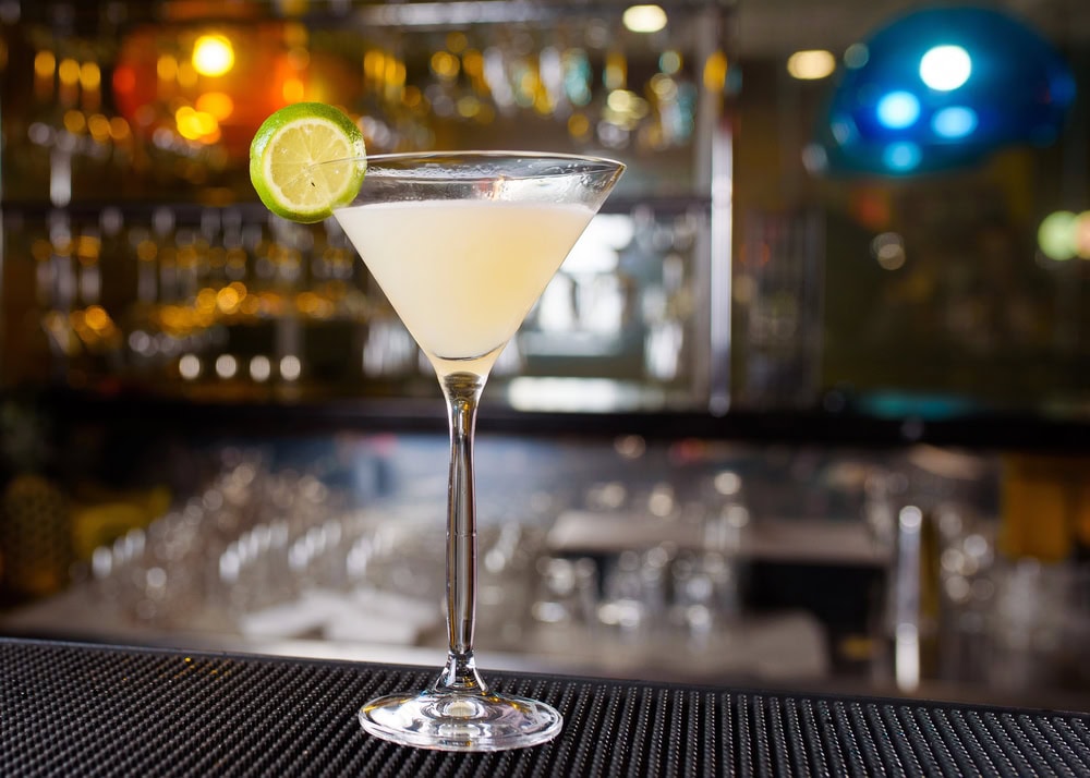 A cocktail in a martini glass garnished with a lime wheel sits on a bar counter, with glassware and bottles blurred in the background.