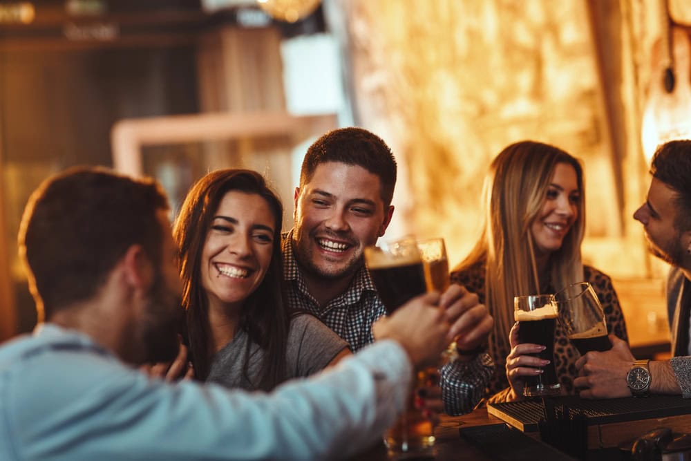 Four people are sitting at a bar, smiling and clinking glasses of dark beer in a casual, warmly lit setting.