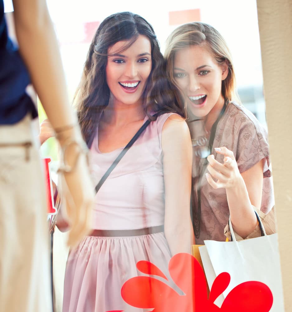 Two women smile and look excitedly at clothing displayed in a store window while holding shopping bags.