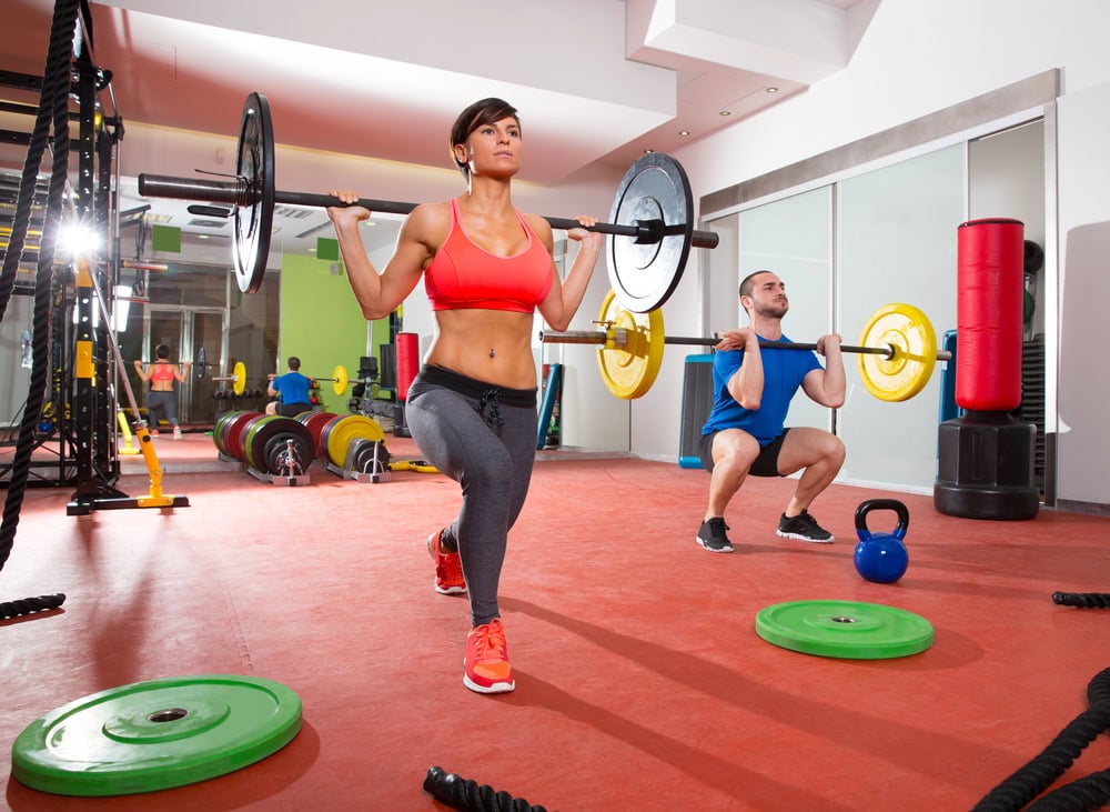 Two people exercise in a gym; a woman does a barbell lunge in the foreground, while a man performs a barbell squat in the background. Gym equipment is visible around them.