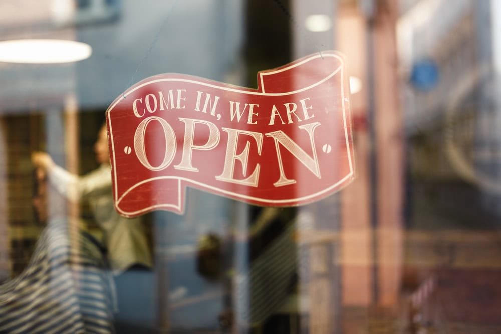 Red and white sign with the text "Come in, we are open" displayed on a glass door or window of a storefront.