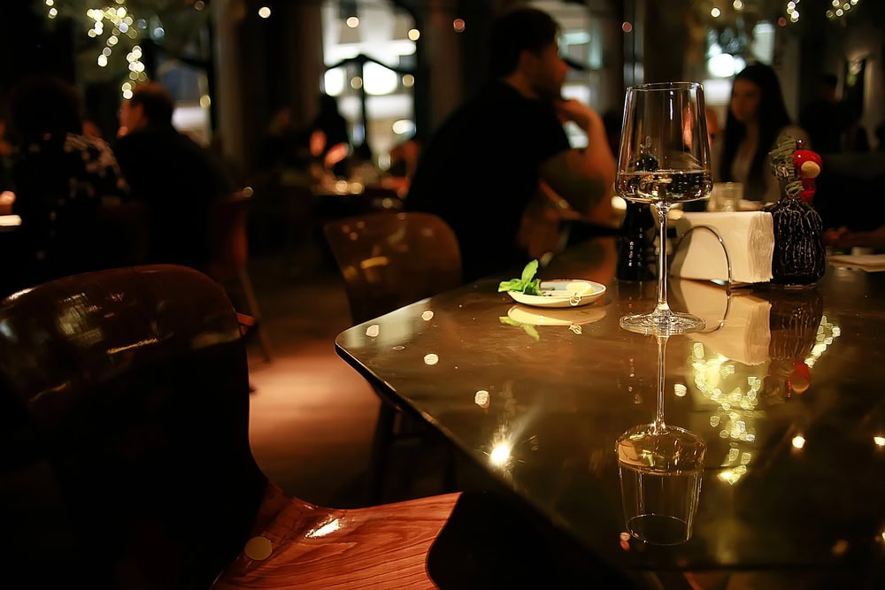 A dimly lit restaurant with people dining in the background, a glass of wine and a small plate with garnish on a reflective table in the foreground.
