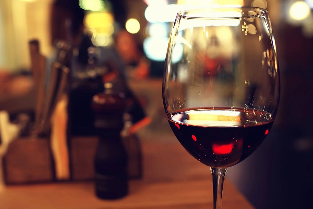 A glass of red wine sits on a table in a dimly lit restaurant with blurred background objects and lights.