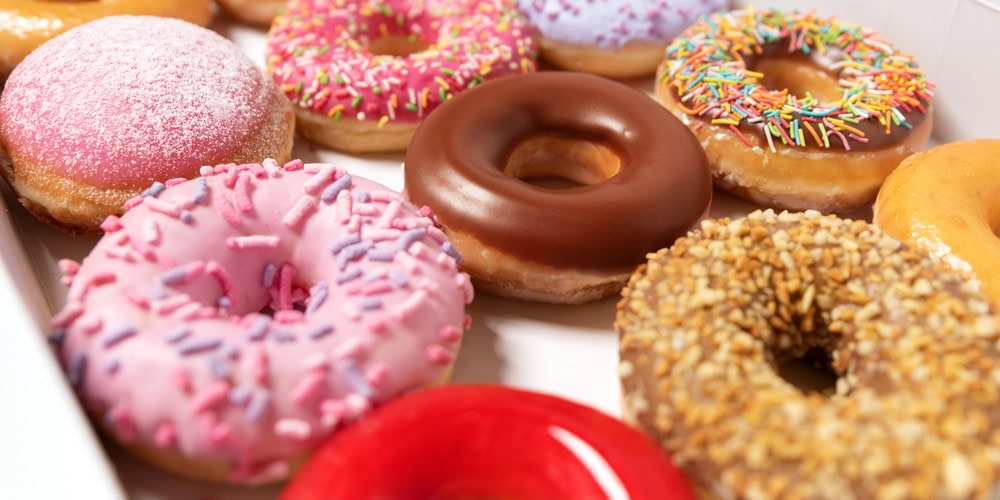 Assorted donuts in a box, including ones with pink icing and sprinkles, chocolate glaze, powdered sugar, crushed nuts, and classic glazed varieties.