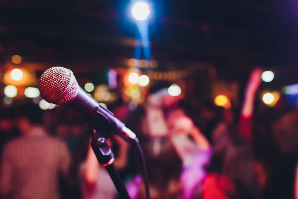 Close-up of a microphone on a stand with colorful stage lights and a blurred crowd in the background, suggesting a live performance or event.