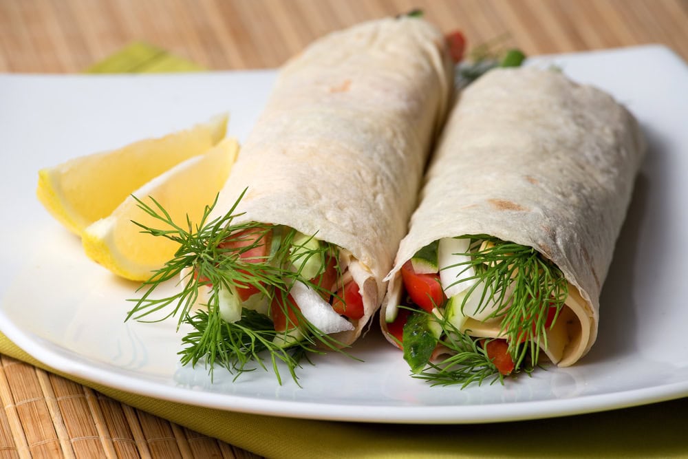 Two vegetable wraps with fresh herbs and sliced vegetables on a white plate, served with two lemon wedges.