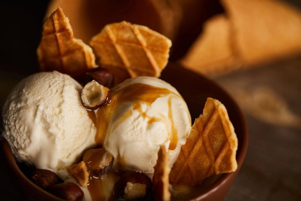 Two scoops of vanilla ice cream in a bowl topped with caramel sauce, hazelnuts, and pieces of waffle cone, with an empty cone in the background.
