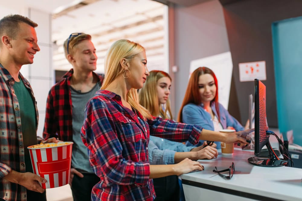 A group of people stand at a counter, with one woman using a touchscreen while others watch. One person holds a bucket of popcorn.