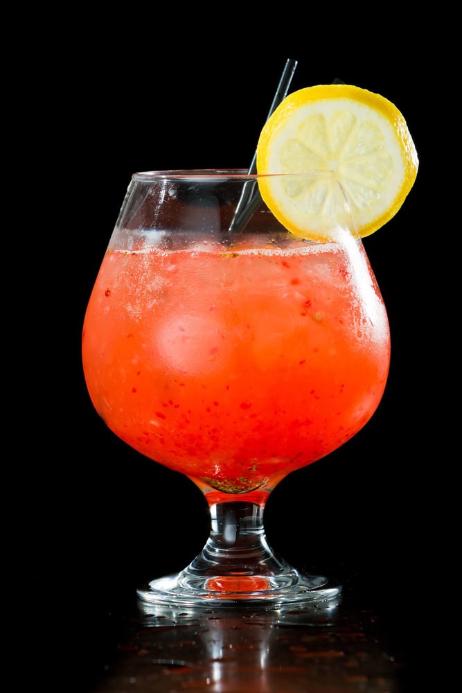 A red cocktail with ice is served in a round glass, garnished with a lemon slice and a black straw, against a black background.