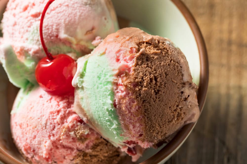 A bowl containing scoops of spumoni ice cream in pink, green, and brown colors, topped with a red cherry.