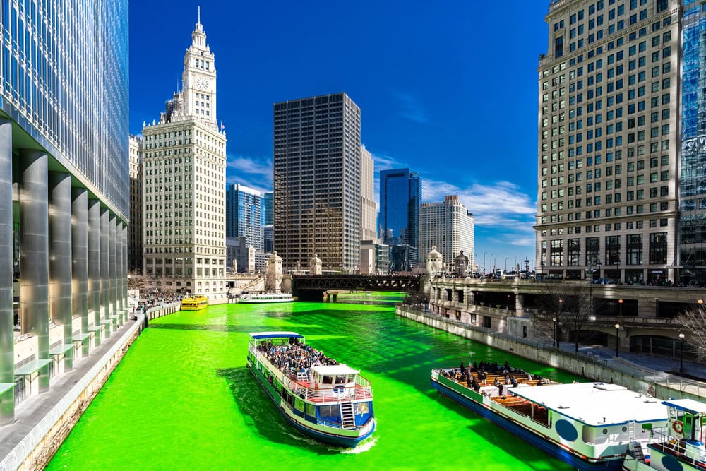 Tour boats travel on the Chicago River, which has been dyed bright green, with tall buildings and a clear blue sky in the background.