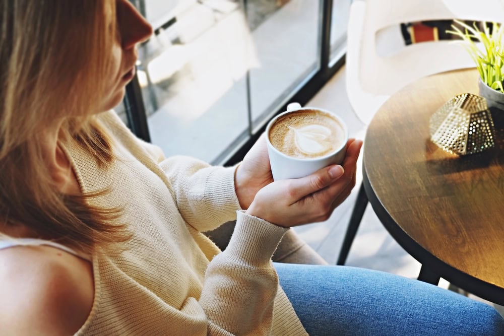 A woman in a cream sweater and jeans sits by a table, holding a cup of coffee with latte art, near a window with natural light.