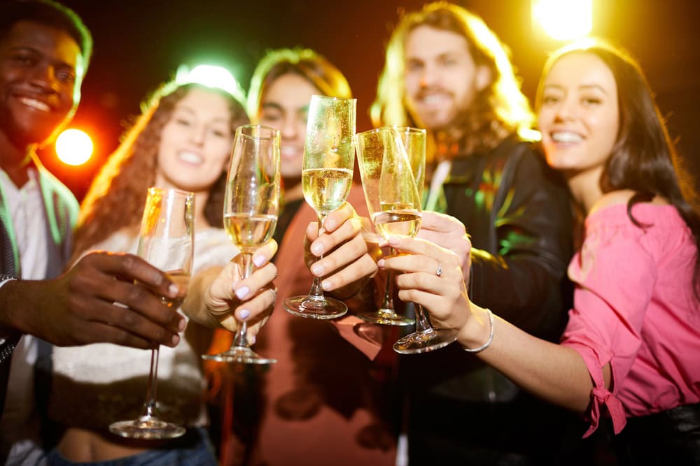 Five people holding champagne glasses, smiling, and making a toast at a party with bright lights in the background.
