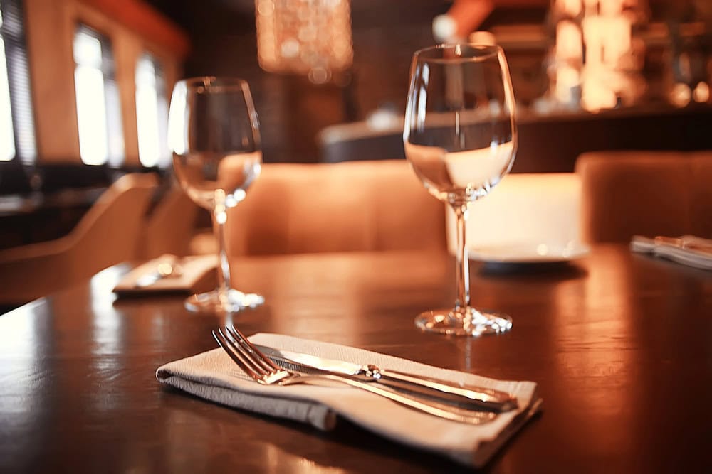 A close-up of a restaurant table set with two empty wine glasses, plates, and cutlery on a napkin, with warm ambient lighting in the background.