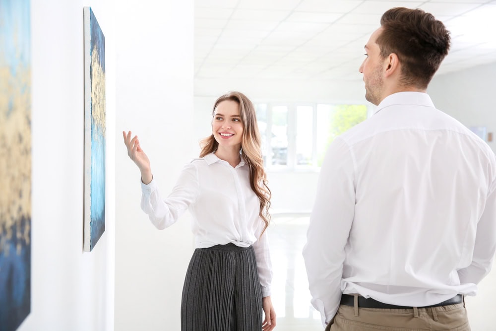 A woman points at a painting on a white wall while talking to a man in a brightly lit gallery space.