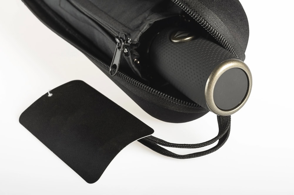 A black cylindrical object partially inside a zippered black case, with a blank black tag attached to the case, on a white background.