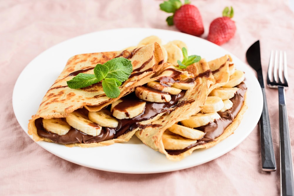 Two folded crepes filled with banana slices and chocolate sauce, garnished with mint leaves, served on a white plate with strawberries in the background.