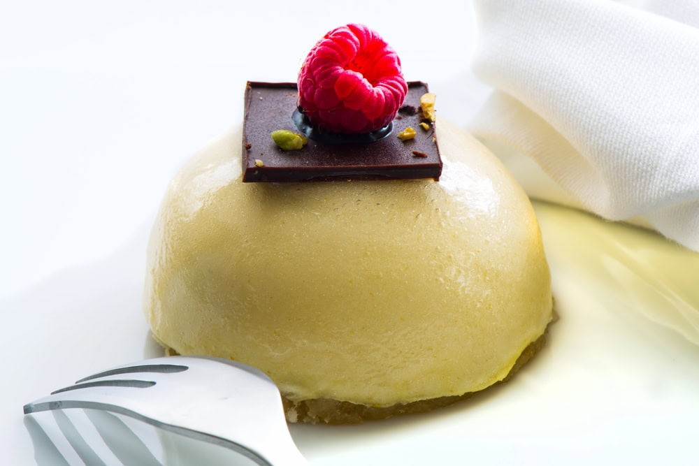 A dome-shaped dessert topped with a square of dark chocolate and a raspberry, placed on a white plate next to a fork and a folded white napkin.