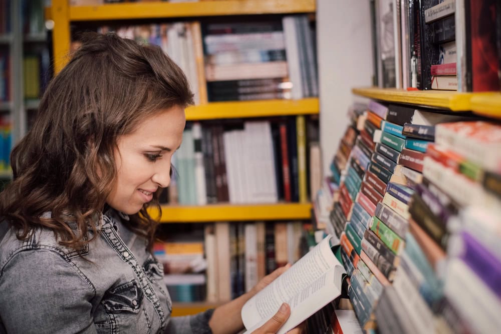 A woman wearing a denim jacket reads a book while standing next to shelves filled with books in a library or bookstore.