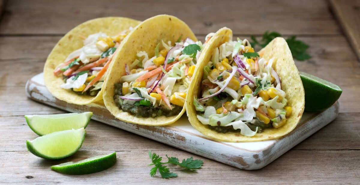 Three tacos filled with mixed vegetables and slaw are arranged on a white board with lime wedges and cilantro on a wooden surface.