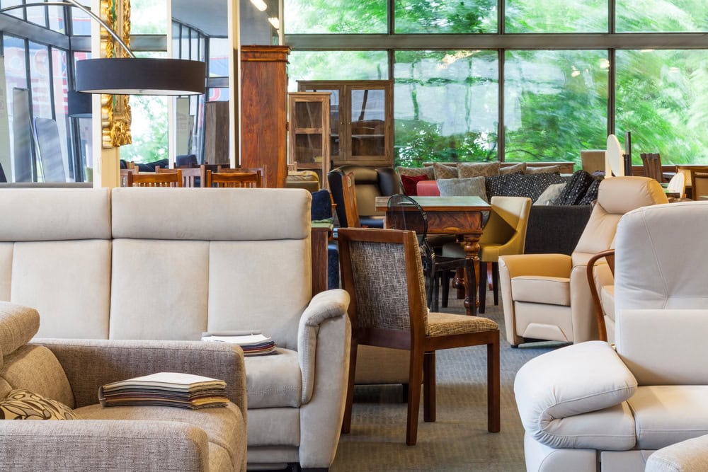 Showroom with various sofas, armchairs, and dining sets displayed near large windows with outdoor greenery visible in the background.