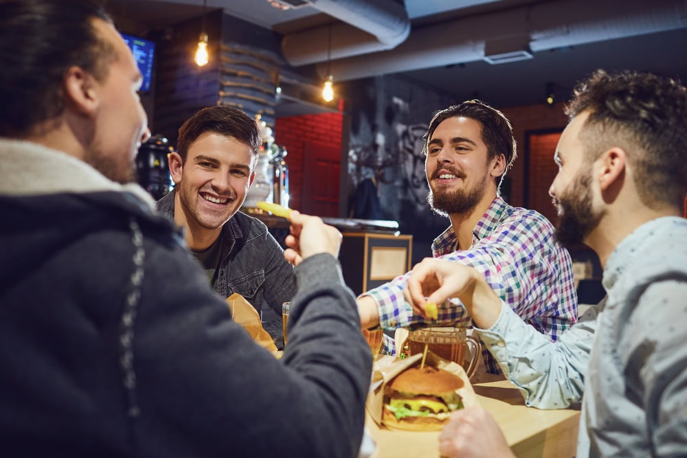 Four men sit at a bar table, talking and laughing, with burgers and drinks in front of them under warm lighting.