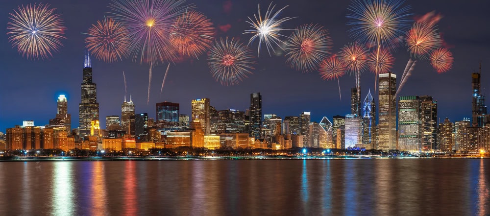 Fireworks light up the night sky above a city skyline with tall buildings, reflected in the water below.