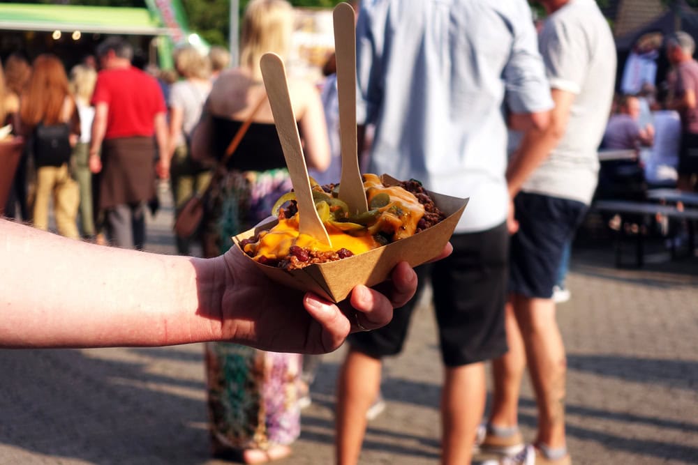 A hand holds a tray of nachos topped with melted cheese and jalapeños, with two wooden forks, at an outdoor event with people in the background.