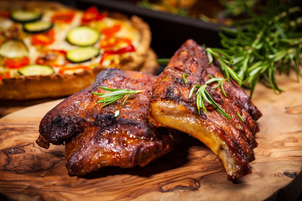 Two pieces of grilled ribs garnished with rosemary on a wooden board, with a vegetable tart in the background.