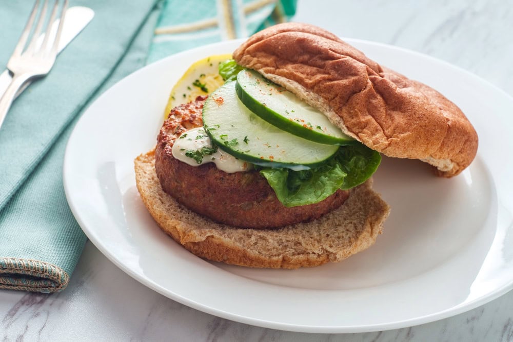 A veggie burger with lettuce, cucumber slices, and sauce on a whole wheat bun sits on a white plate beside a teal napkin with utensils.
