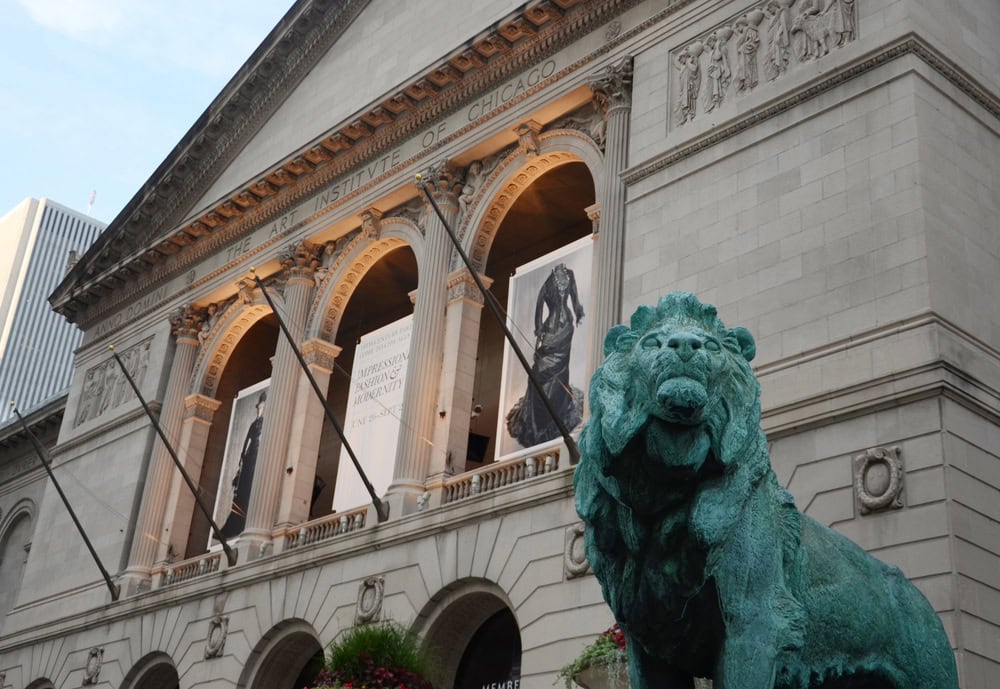 The exterior of the Art Institute of Chicago with its bronze lion statue in front and two art banners hanging from the building's facade.